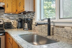 A kitchen with a granite countertop, a stainless steel sink, wooden cabinets, a coffee maker, and a window with a view of trees.