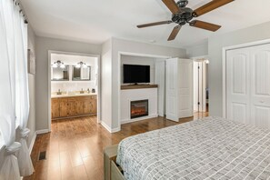 Un dormitorio con una cama grande, ventilador de techo, chimenea, televisor y un baño con tocador y espejo.