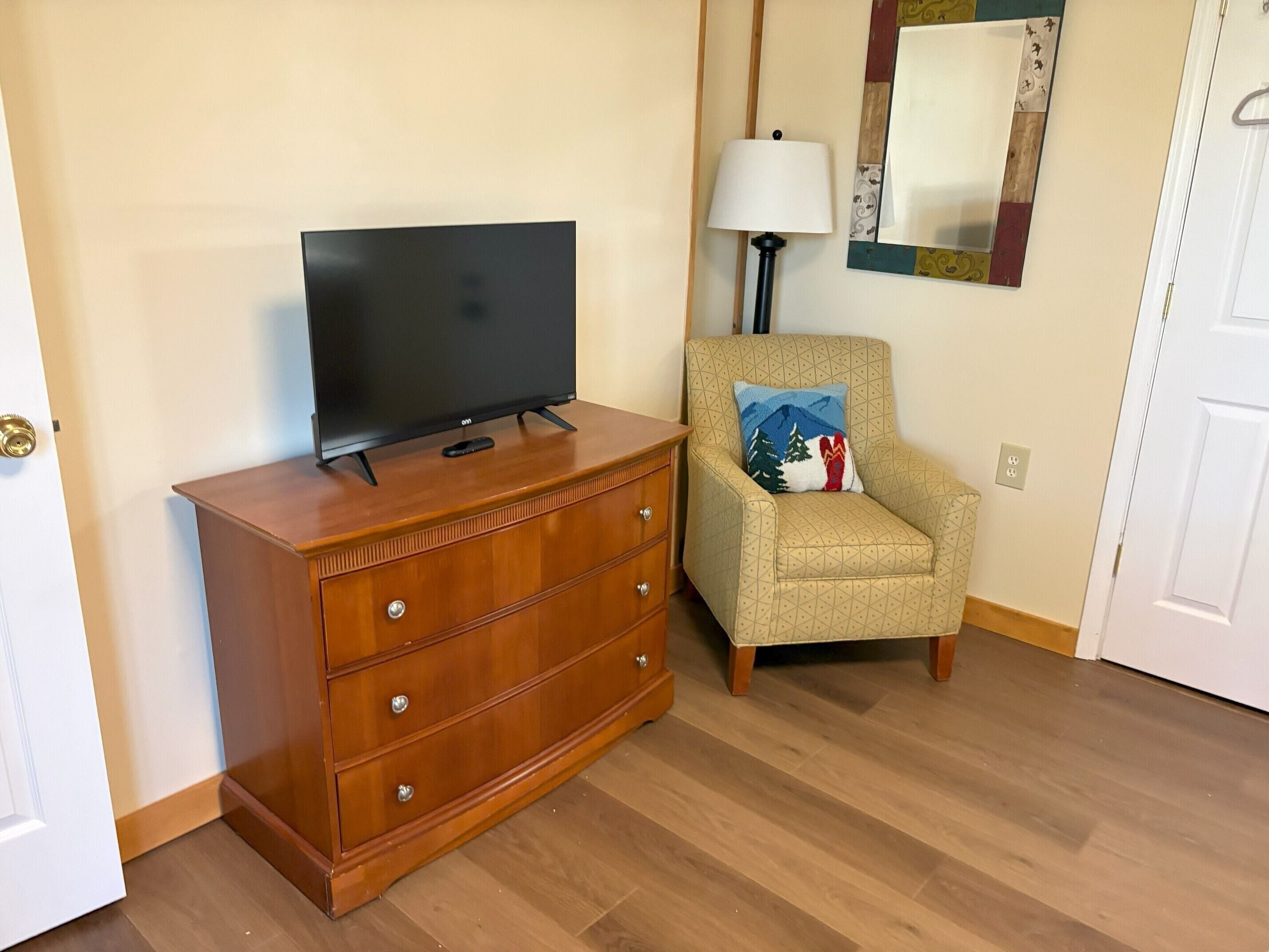 Une pièce comprenant une commode en bois, un téléviseur, une lampe, une chaise avec un coussin à motifs et un miroir au mur.