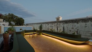 Uma área externa com assentos, incluindo um banco curvo e plantas em vasos, com uma parede texturizada e uma estrutura em forma de cúpula ao fundo.