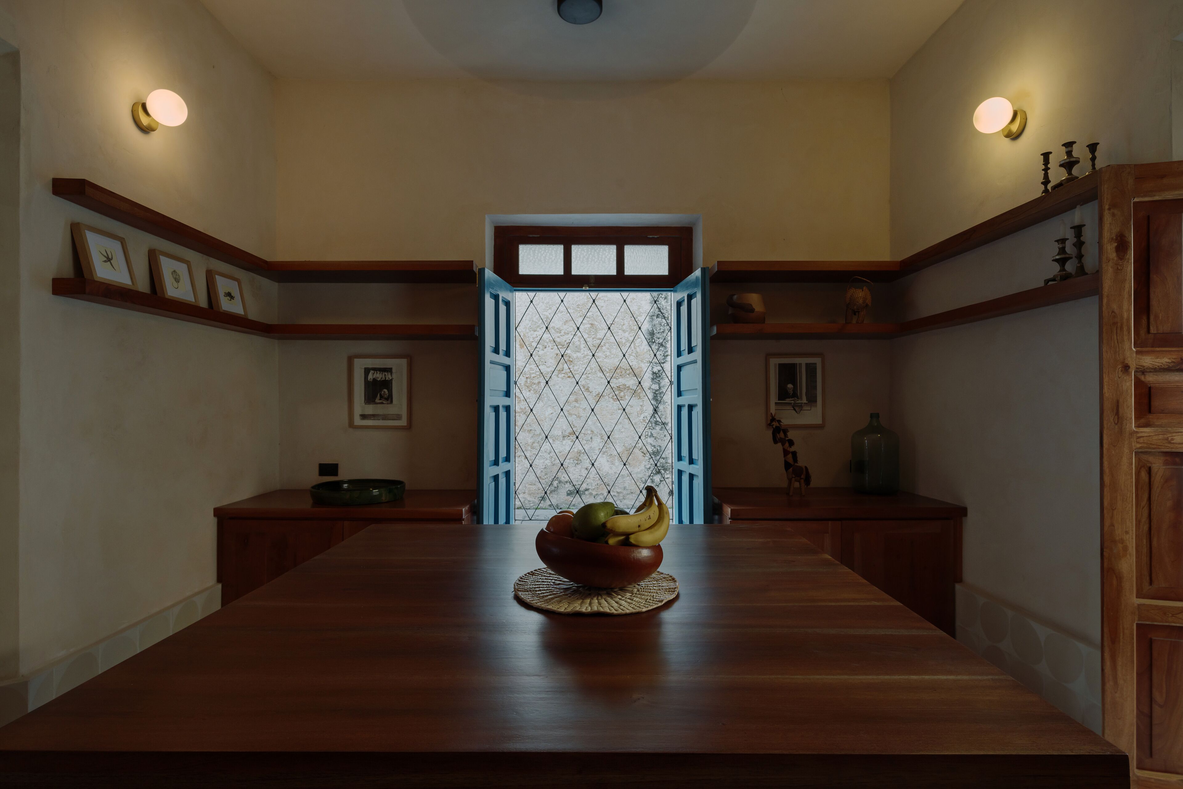 A dining area with a long wooden table, a bowl of fruit, and a patterned window with blue shutters.