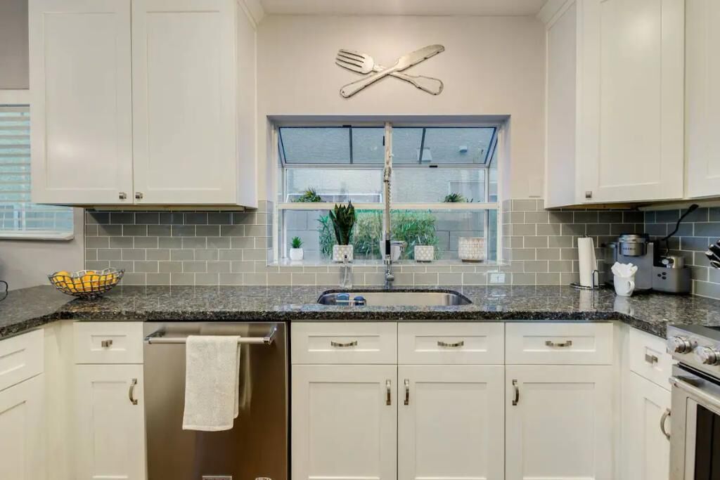 Una cocina con armarios blancos, encimera de granito, ventana con plantas y un adorno de utensilios sobre el fregadero.