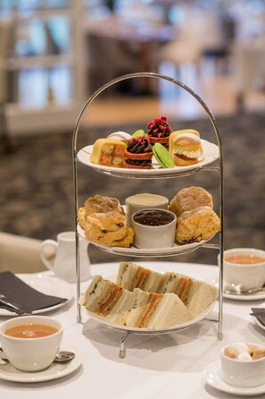 Una bandeja de tres niveles con una variedad de pasteles y sándwiches, acompañada de tazas de té y café, sobre una mesa en un entorno hotelero.