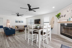 Ein modernes Wohnzimmer mit Essbereich, zwei Deckenventilatoren, einem Kamin und einer Surfbrett-Dekoration.