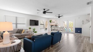 Un salon spacieux avec un canapé, des fauteuils bleus, une table basse et un téléviseur. On y trouve également des ventilateurs de plafond, un coin repas et une vue dégagée grâce à des portes vitrées.