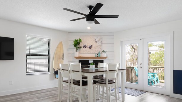 Un coin repas avec une table, des chaises, un ventilateur de plafond, une planche de surf et une vue à travers la porte vitrée.