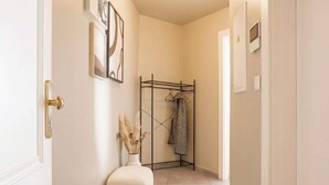 A hallway with a white door, a white wall with framed artwork, a black metal rack with clothes, a white pouf, and a vase with dried plants. - TurnKey | Angelo Roma Studio – Cozy Balcony (Praha 2)