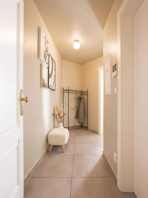 A hallway with a white door, a white wall with framed artwork, a black metal rack with clothes, a white pouf, and a vase with dried plants. - TurnKey | Angelo Roma Studio – Cozy Balcony (Praha 2)