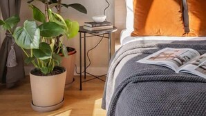 A neatly arranged bedroom with a bed, a bedside table with a lamp, a potted plant, and a framed picture on the wall. - TurnKey | Angelo Roma Studio – Cozy Balcony (Praha 2)