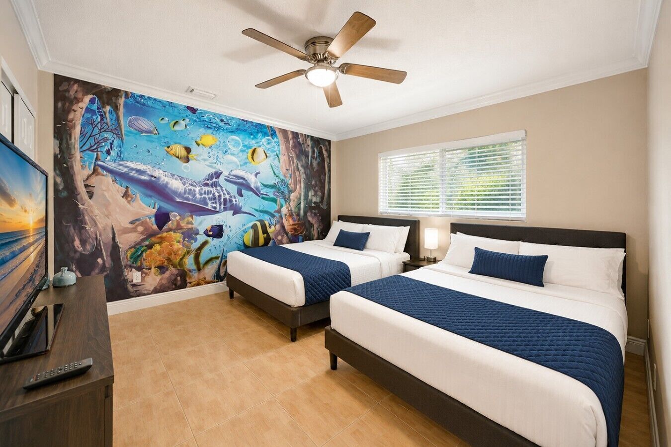 A hotel room with two beds, a ceiling fan, a mural of marine life, and a window with blinds.