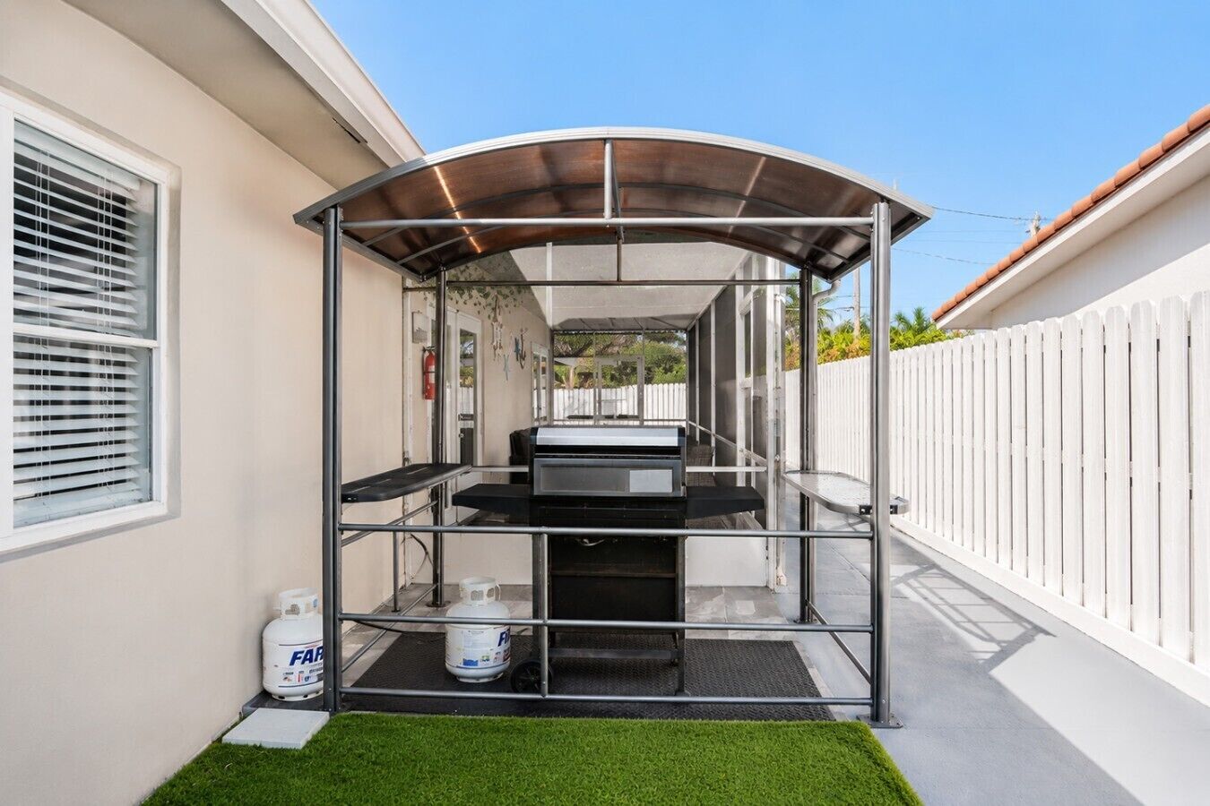 A covered outdoor kitchen area with a grill, a gas tank, and a white fence in the background.