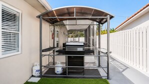 Eine überdachte Außenküche mit Grill, Gasflasche und weißem Zaun im Hintergrund.