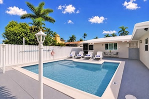 A rectangular swimming pool with a white fence, lounge chairs, and a small table.