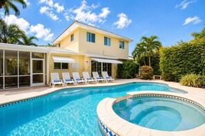 Une piscine avec des chaises longues, un patio couvert et un bâtiment à deux étages surmonté d’un toit en tuiles.
