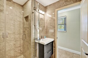 Un baño moderno con ducha de vidrio, un lavamanos blanco con gabinete oscuro y piso de baldosas color beige.