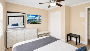 Habitación de hotel con cama, escritorio, silla, ventilador de techo, puerta blanca y un cuadro de paisaje urbano en la pared.