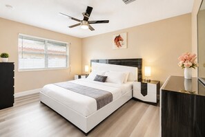 Ein Schlafzimmer mit einem Bett, Nachttischen, einem Deckenventilator, einem Fenster mit Jalousien und einem Gemälde an der Wand.