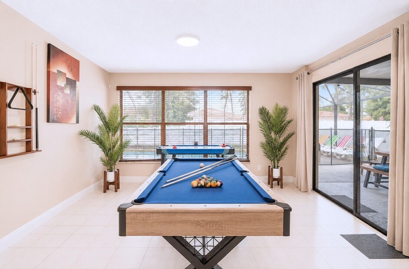 Une table de billard dans une pièce dotée de grandes fenêtres, d’une porte donnant sur l’extérieur et de plantes en pot.