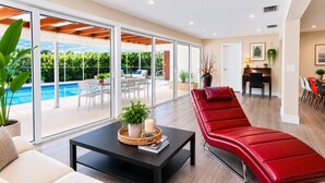 Una sala moderna con un sillón rojo, una mesa de centro negra con objetos decorativos, un sofá blanco y vistas a un área de piscina exterior a través de amplias puertas de vidrio.