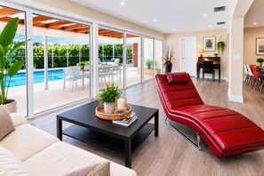 Una sala moderna con un sillón rojo, una mesa de centro negra con objetos decorativos, un sofá blanco y vistas a un área de piscina exterior a través de amplias puertas de vidrio.