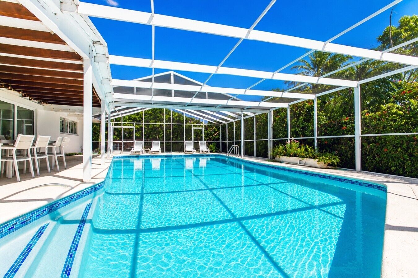 A clear blue swimming pool with a white roof structure, surrounded by white lounge chairs and a patio area.