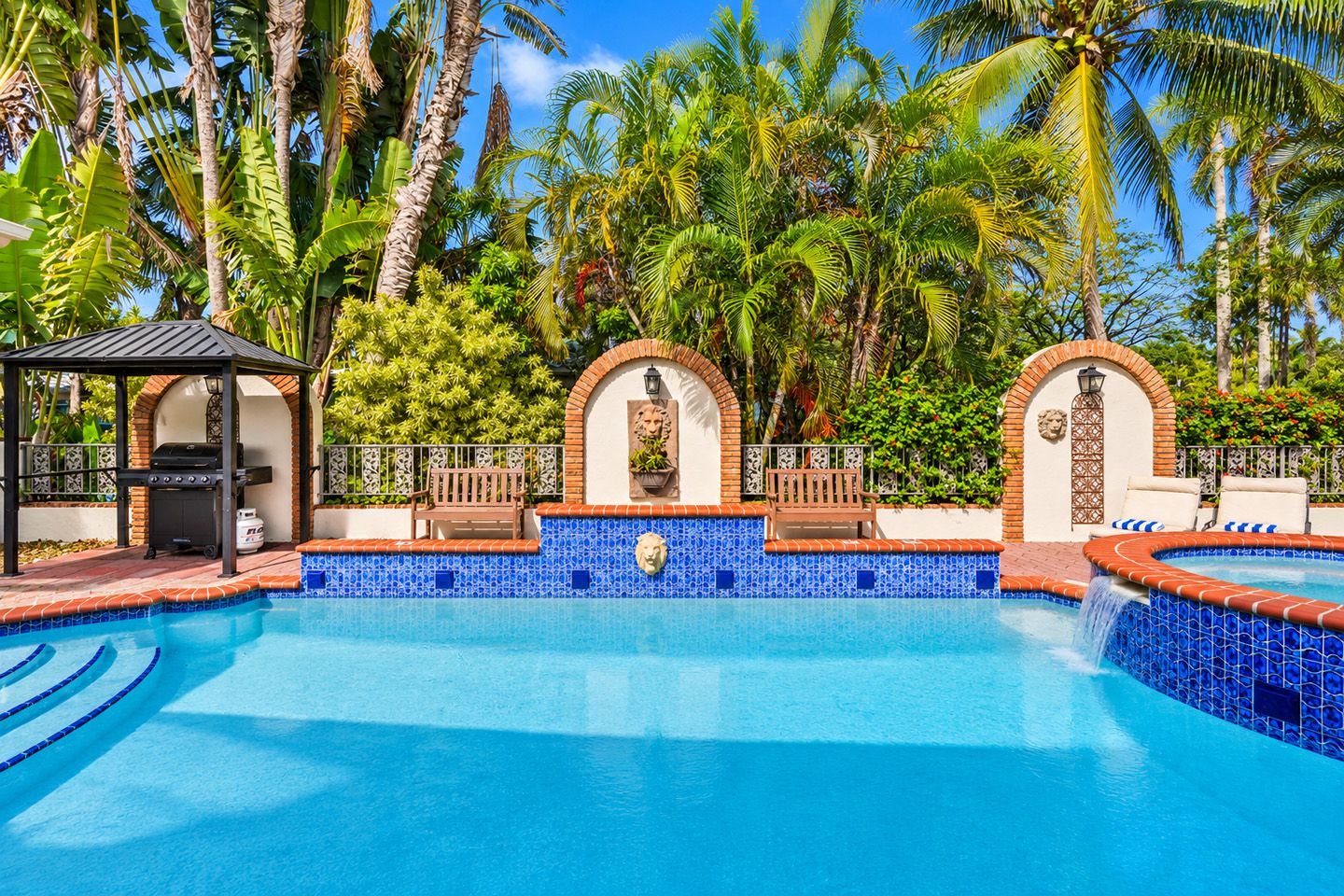Ein Swimmingpool mit einer gefliesten Kante, umgeben von Palmen und einem Pavillon mit Grill.