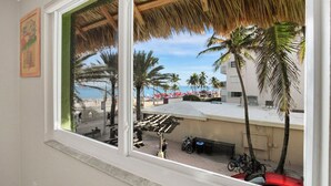 A window view of a beach with palm trees, a thatched roof structure, and a building with a sign that reads "COSTA DEL MAR". - Beach Condo - Hollywood Beachfront & Broadwalk (Hollywood)