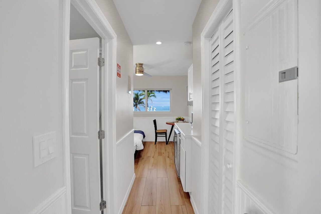 A narrow hallway with white walls and wooden flooring leading to a small kitchen area with a window overlooking the ocean.
