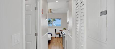 A narrow hallway with white walls and wooden flooring leading to a small kitchen area with a window overlooking the ocean.