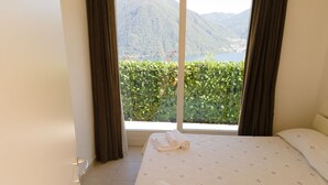 A hotel room with a bed, a window with curtains, and a view of a mountain and lake. - Villa Rosa - My Home in Como (Muronico)