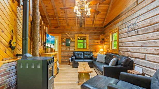 A log cabin interior with a fireplace, leather sofas, a wooden table, and a ceiling fan.