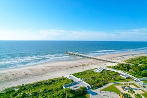 Vista aérea de uma praia com um píer que se estende para o oceano, extensa faixa de areia e área de parque com caminhos e vegetação.