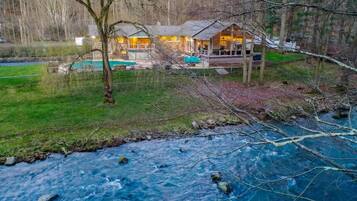 A cabin with a deck overlooking a river, surrounded by trees and a grassy area.