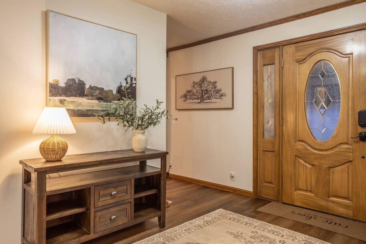 A wooden console table with a lamp and a vase of plants, a framed landscape painting, and a wooden door with a glass panel.