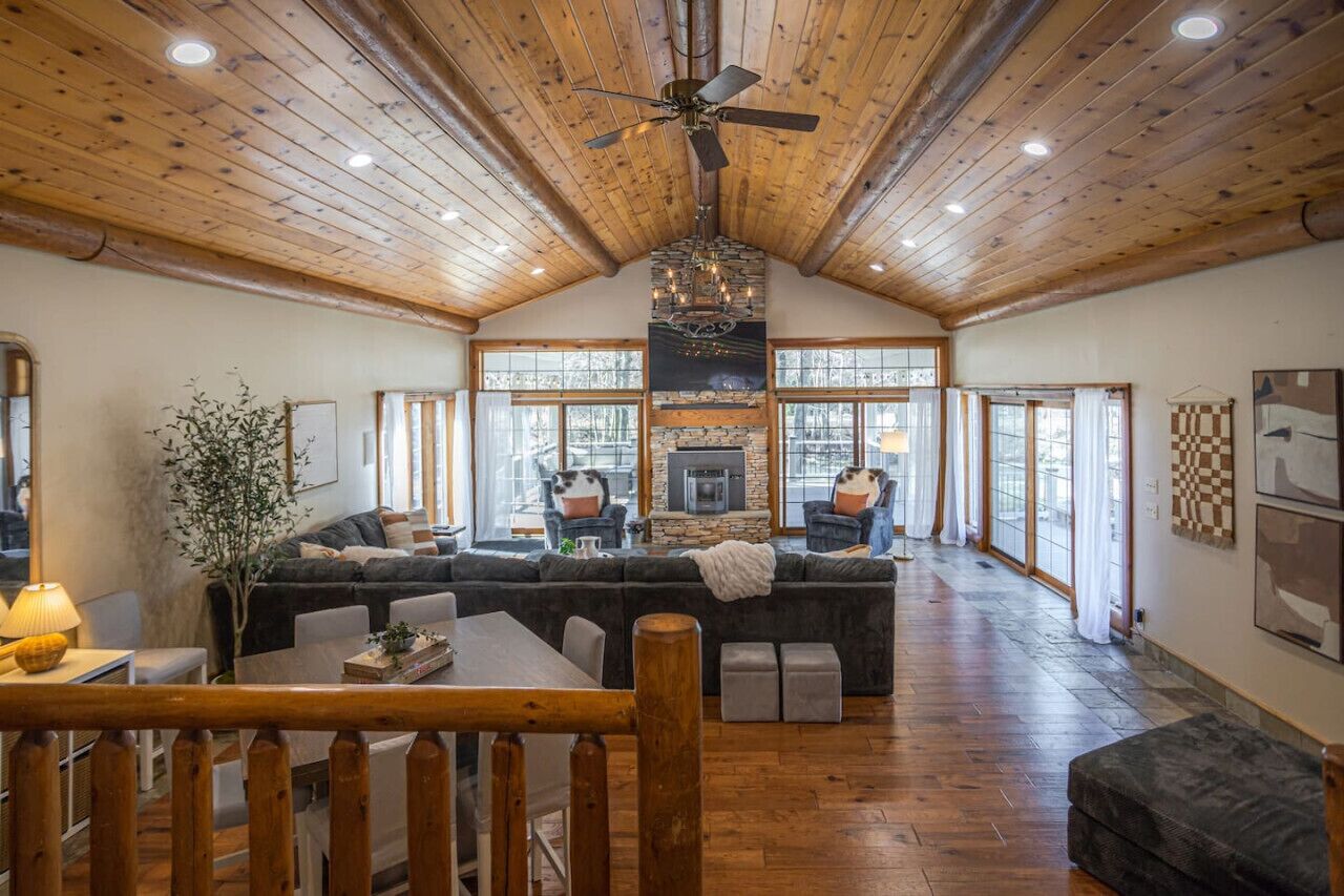 A spacious living area with a large fireplace, wooden ceiling, and a view through large windows.
