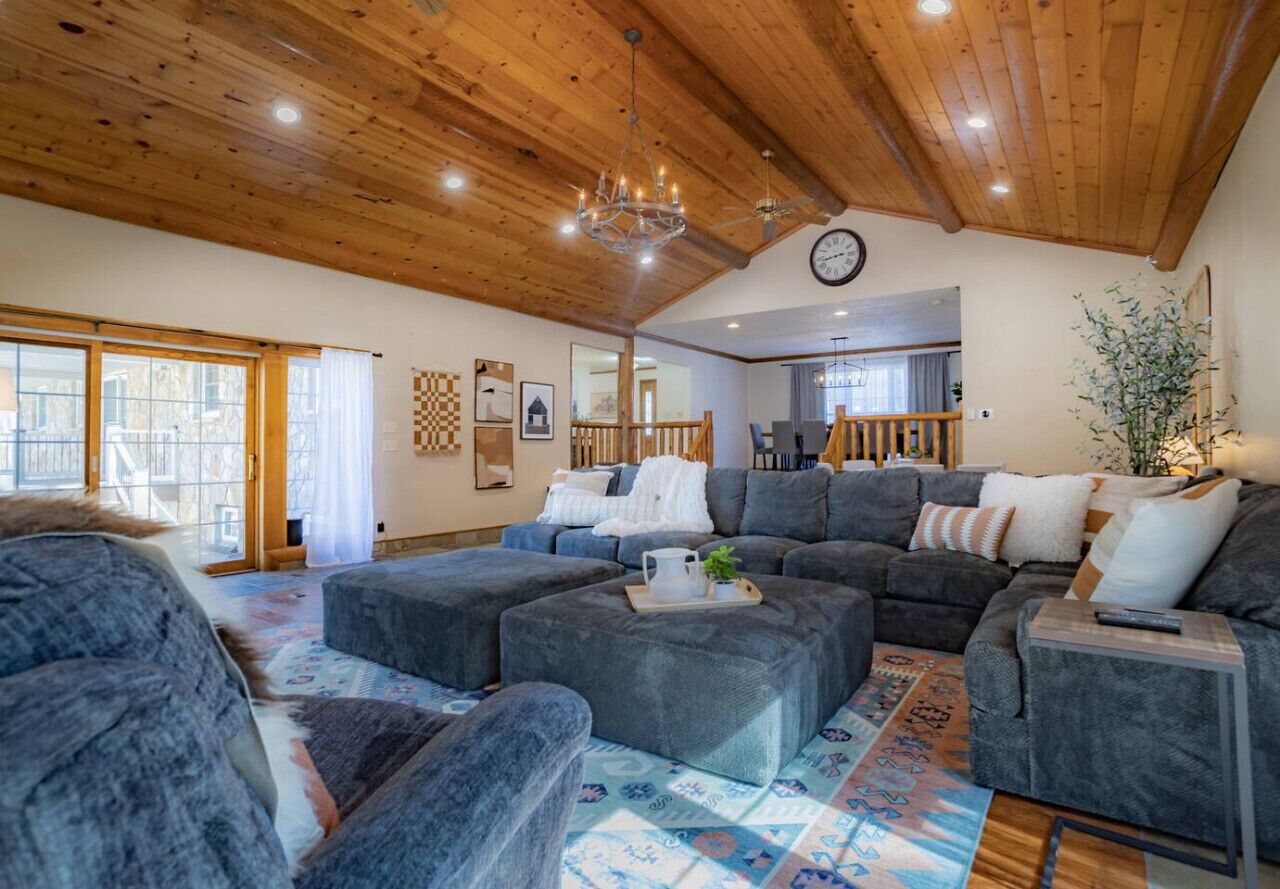 Ein geräumiges Wohnzimmer mit einem großen, grauen Ecksofa, Hocker und einer Holzbalkendecke.