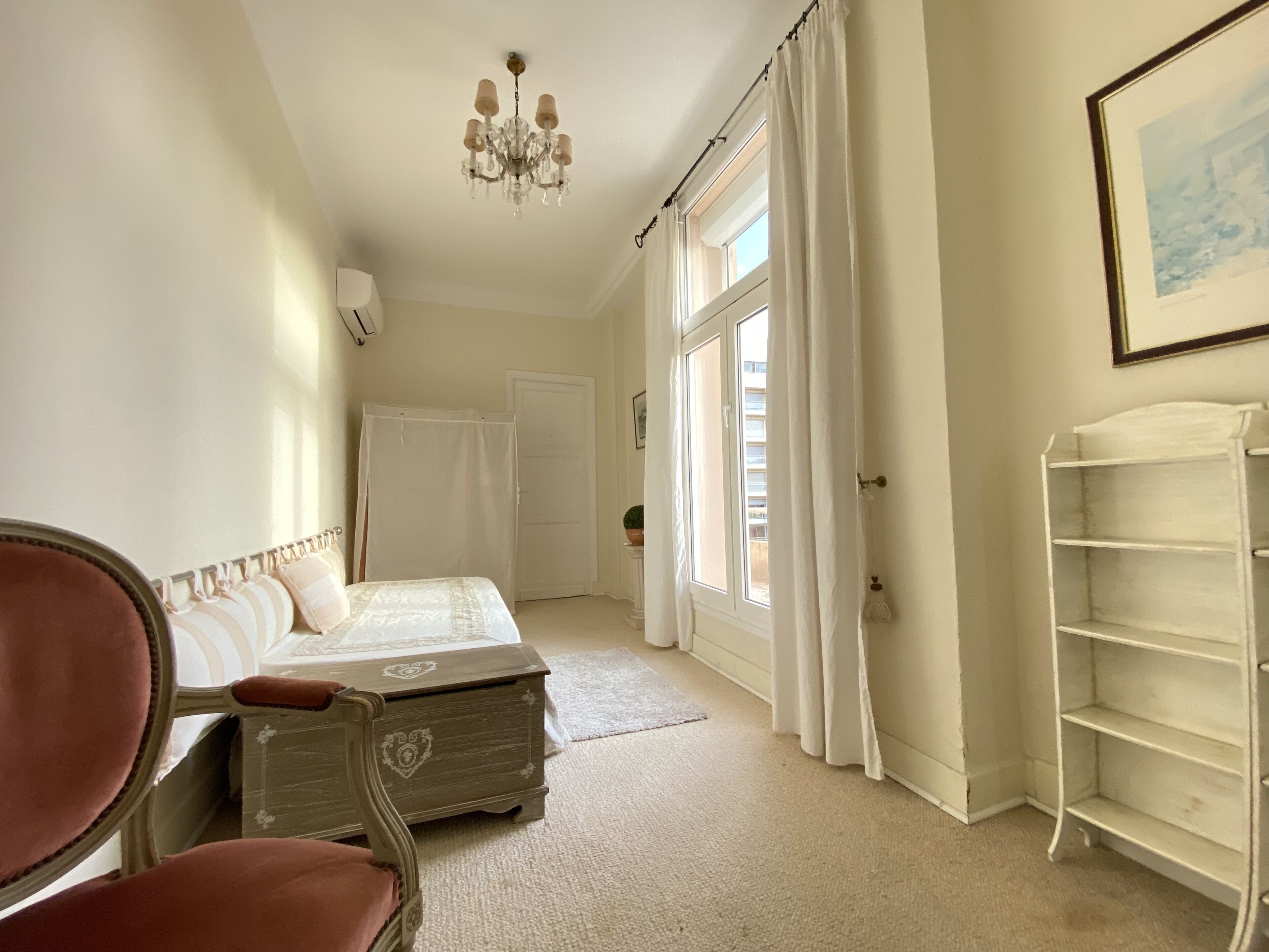 A bedroom with a bed, a chair, a chest of drawers, a chandelier, a window with curtains, and a framed picture on the wall.