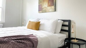 Una cama bien hecha con ropa blanca y una manta decorativa estampada, una sola almohada de color mostaza, y un cuadro abstracto moderno en la pared.