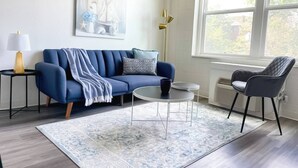 Una sala de estar moderna con un sofá azul, una mesa de centro redonda y un sillón gris. Hay una alfombra estampada en el suelo, una lámpara en una mesita auxiliar y un cuadro en la pared.