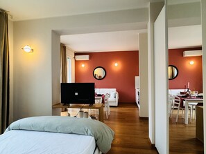 Ett hotellrum med en säng, en TV på ett stativ, ett skrivbord med en stol, ett matbord med stolar och en spegel på väggen.