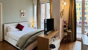 A modern hotel room with a bed, TV, bedside table, and a window with a city view. - The Little Beauty Studio (Pisa)