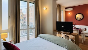 A hotel room with a bed, a television, a desk, and a view of buildings outside the window. - The Little Beauty Studio (Pisa)