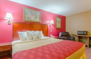 A hotel room with a large bed, two bedside lamps, a wooden headboard, a chair, a desk with a radio, and two framed pictures on the wall.