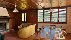 A cozy living room with a fireplace, a yellow sofa, a dining table with chairs, and large windows with blinds. - Monte Bar Old Cottage (Lugano)