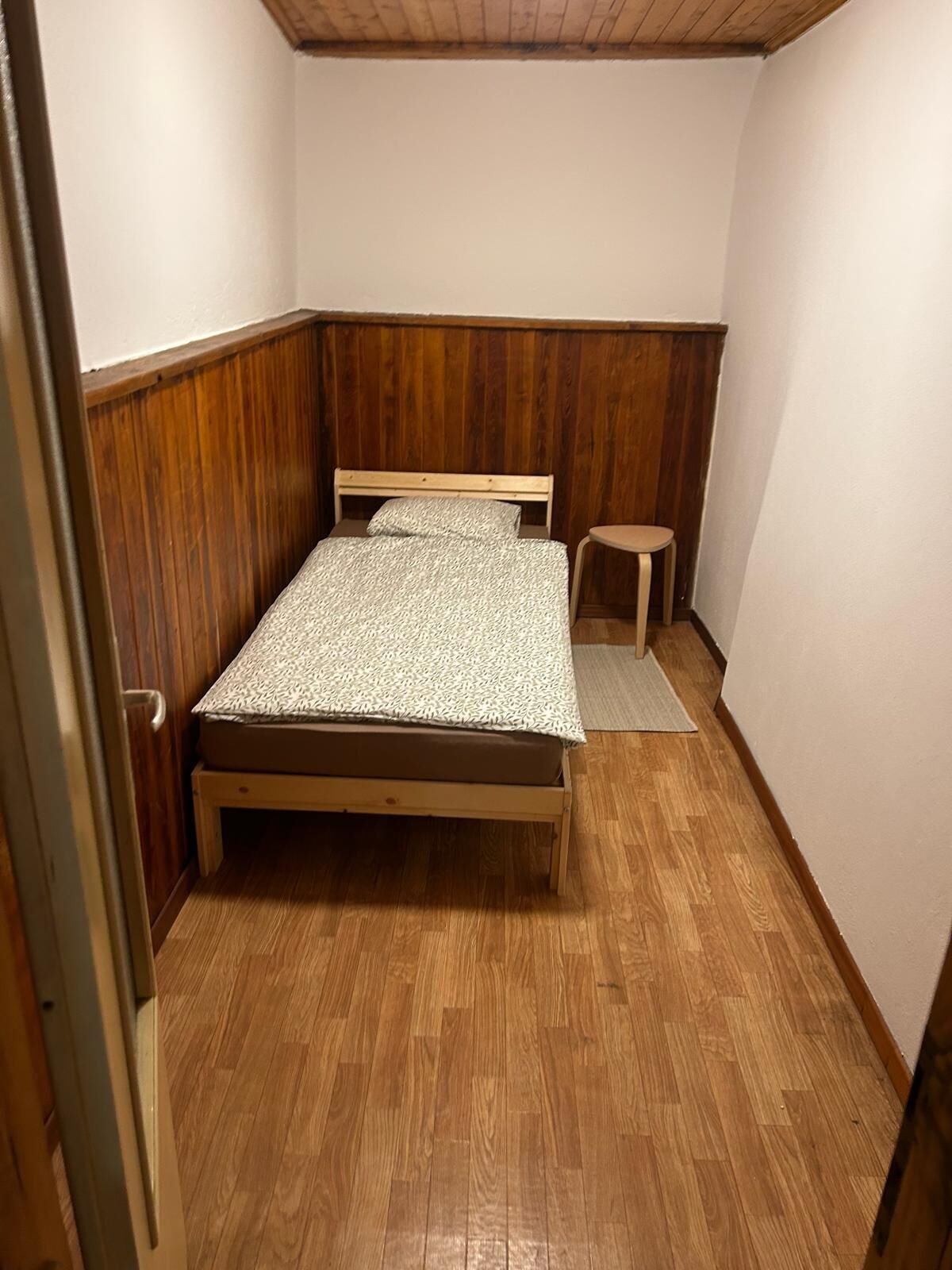 A small, cozy bedroom with a wooden bed, a small stool, and a wooden floor.
