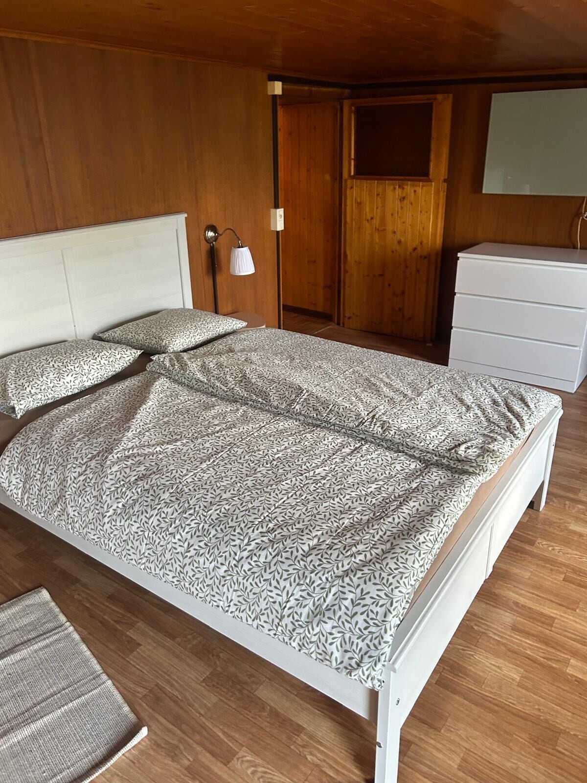 A bedroom with a bed, a nightstand, a lamp, a dresser, and wooden walls.