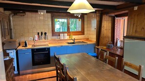 A compact kitchen with a wooden table and chairs, a built-in oven, a sink, and a window with a view of greenery. - Monte Bar Old Cottage (Lugano)