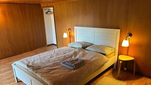 A neatly made bed with patterned bedding, two bedside lamps, a small stool, and a woven mat on a wooden floor. - Monte Bar Old Cottage (Lugano)