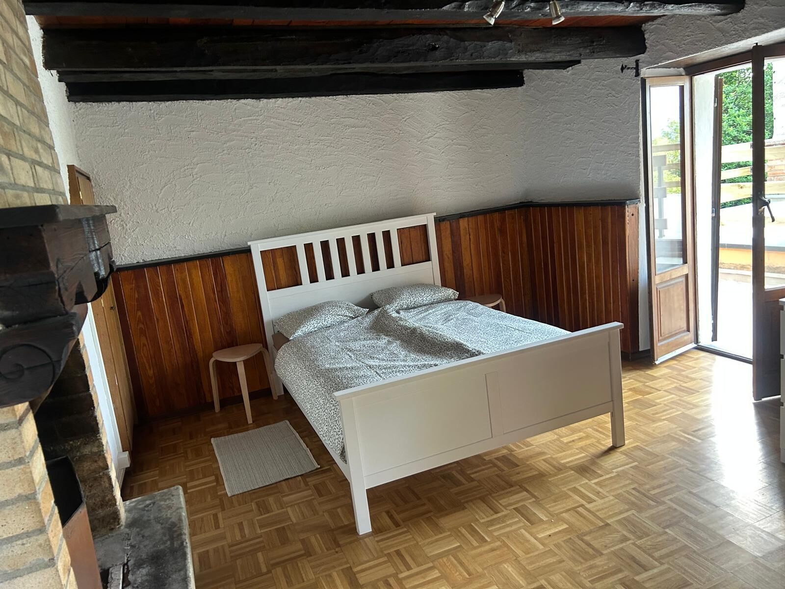 Quarto com piso de madeira, cama branca e um banquinho pequeno.