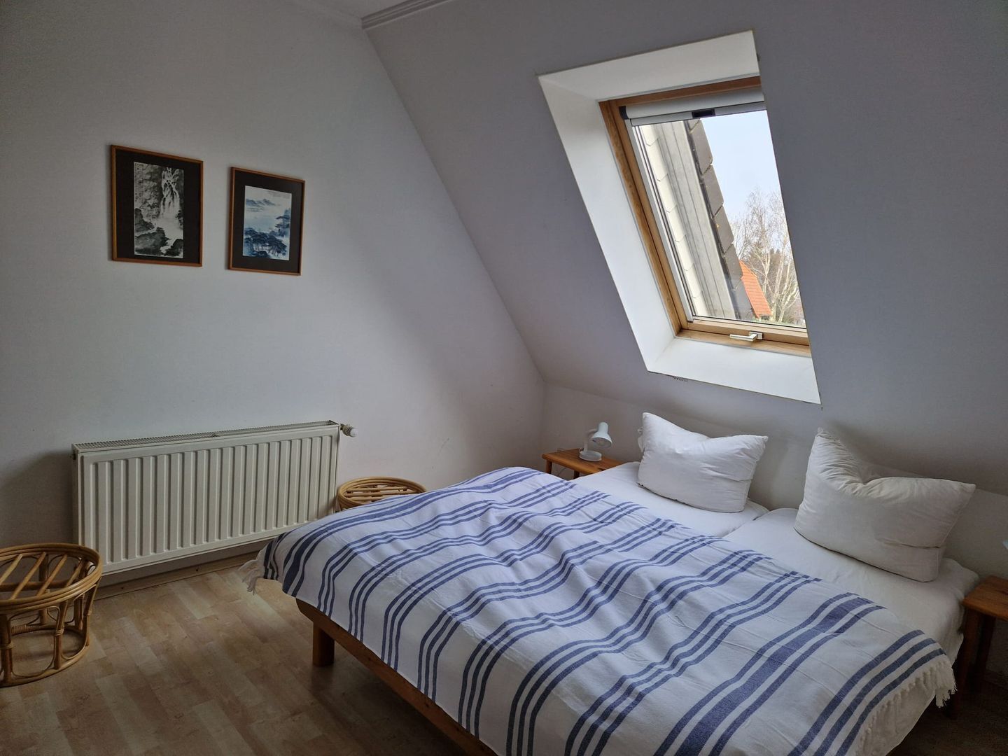 Un dormitorio con techo inclinado, una cama con ropa de cama azul y blanca a rayas, dos cuadros en la pared, un radiador y una ventana con vistas al exterior.